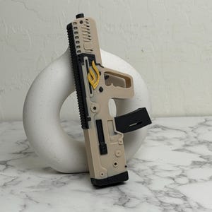 Può includere: Una pistola giocattolo beige e nera con un'emblema dorato. La pistola giocattolo ha un caricatore, un'impugnatura e un mirino. La pistola è in plastica e presenta un design dettagliato.