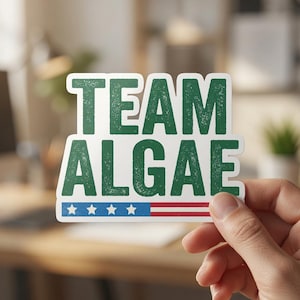 May include: A die-cut sticker featuring the text TEAM ALGAE in a distressed dark green font. Below the text is a horizontal graphic element displaying a blue field with four white stars on the left and red and white horizontal stripes on the right.