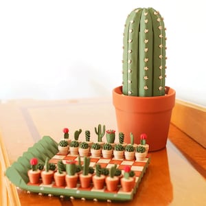 May include: A decorative cactus in a terracotta pot, with a green cactus-shaped chess set. The chess set features miniature cacti in small pots on a checkered board. The large cactus has small pink star-shaped accents.