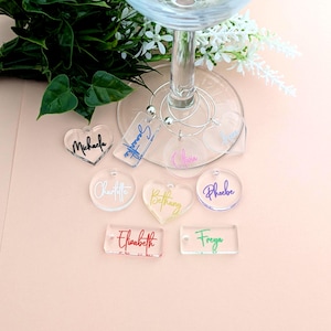 May include: Clear acrylic wine glass charms in various shapes, including hearts and circles, with names written in different colours. The charms are displayed near a wine glass and greenery, ideal for a party or event.