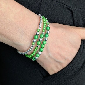 May include: Three beaded bracelets. Two bracelets feature bright green beads, and one bracelet has silver beads. The bracelets are worn on a wrist, and the background is a black garment.