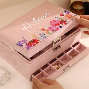 Può includere: Un portagioie in velluto rosa con più scomparti e cassetti. Il coperchio è decorato con un motivo floreale e il nome "Arlene" in corsivo bianco. La scatola è aperta, rivelando l'interno e alcuni gioielli.