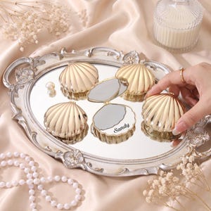 May include: Several gold-colored, shell-shaped compact mirrors with a metallic finish. One mirror is open, revealing a reflective surface and the name Sandy printed in black script. The mirrors are arranged on an oval, ornate silver-toned tray.