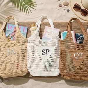 Puede incluir: Tres bolsos de playa de ganchillo en beige, blanco y marr&oacute;n, cada uno con una inicial. Las bolsas est&aacute;n llenas de art&iacute;culos esenciales de playa como revistas, protector solar y toallas. La escena est&aacute; ambientada sobre una superficie de madera con arena y accesorios de playa.