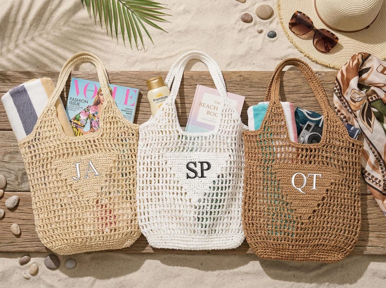 Puede incluir: Tres bolsos de playa de ganchillo en beige, blanco y marr&oacute;n, cada uno con una inicial. Las bolsas est&aacute;n llenas de art&iacute;culos esenciales de playa como revistas, protector solar y toallas. La escena est&aacute; ambientada sobre una superficie de madera con arena y accesorios de playa.