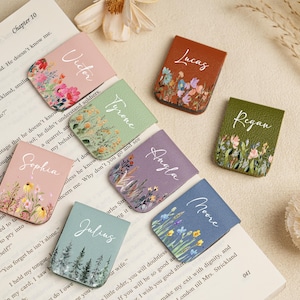 Peut inclure: Ensemble de marque-pages en cuir personnalisés, chacun avec un nom et un motif floral. Les marque-pages sont de différentes couleurs, dont le rose, le marron, le vert et le bleu. Les noms sont écrits en cursive.