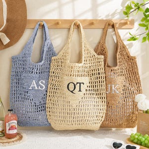 Op de afbeelding: Drie gehaakte draagtassen hangen aan een houten rek. De linker tas is lichtblauw met de letters AS, de middelste tas is crème met de letters QT en de rechter tas is bruin met de letters JK. Elke tas heeft een open geweven ontwerp.