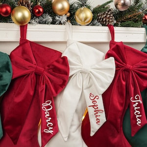 Può includere: Calze di Natale in velluto rosso, bianco e verde, ognuna ornata da un grande fiocco e targhette con nomi personalizzati. Sono appese a un caminetto bianco decorato con ornamenti e verde.