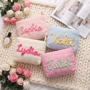 Peut inclure: Quatre pochettes rectangulaires matelassées avec une texture à motif de cœurs, chacune portant un prénom personnalisé en lettres de laine en relief. Les pochettes sont rose clair avec 'Sophia', crème avec 'Lydia', bleu clair avec 'Violet' et rose avec 'Juliet'.