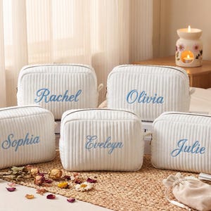 May include: Five white rectangular corduroy cosmetic bags with a zippered closure. Each bag features a unique name embroidered in light blue script: Rachel, Olivia, Sophia, Eveleyn, and Julie. The bags have a vertical ribbed texture.