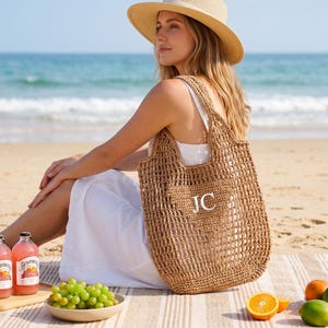 Puede incluir: Un bolso tote tejido y de tejido abierto con las iniciales "JC" en blanco es sostenido por una persona en una playa. El bolso es de color marr&oacute;n natural. La persona lleva un vestido blanco y un sombrero de paja. Hay bebidas y fruta cerca.