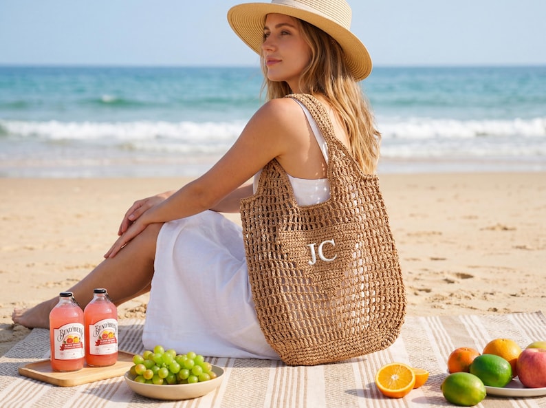 Puede incluir: Un bolso tote tejido y de tejido abierto con las iniciales "JC" en blanco es sostenido por una persona en una playa. El bolso es de color marr&oacute;n natural. La persona lleva un vestido blanco y un sombrero de paja. Hay bebidas y fruta cerca.