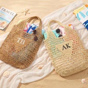 Puede incluir: Dos bolsas de playa tejidas con asas, una con "TB" y la otra con "AK" bordado en la parte delantera. Cada bolsa contiene una toalla enrollada y gafas de sol. Se ve una revista con la palabra "TRAVEL", junto con conchas marinas y una toalla de playa doblada.