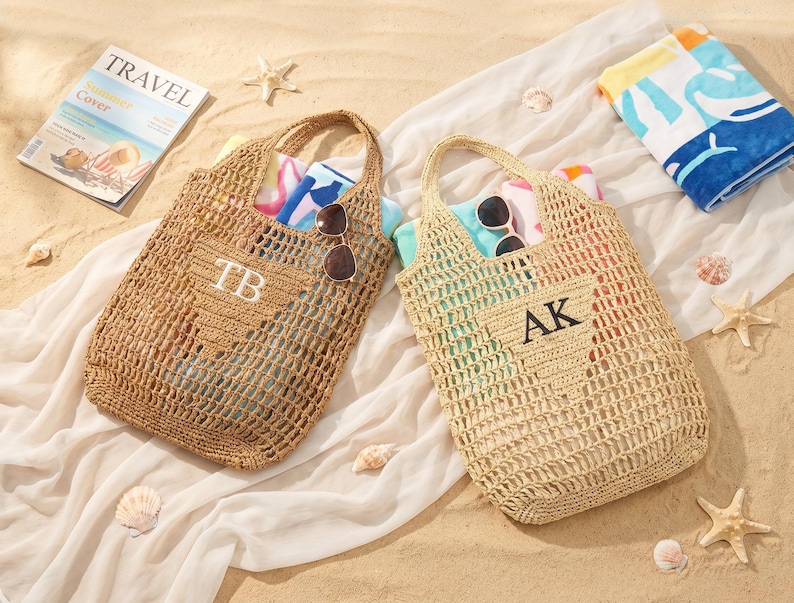 Puede incluir: Dos bolsas de playa tejidas con asas, una con "TB" y la otra con "AK" bordado en la parte delantera. Cada bolsa contiene una toalla enrollada y gafas de sol. Se ve una revista con la palabra "TRAVEL", junto con conchas marinas y una toalla de playa doblada.