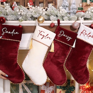 Puede incluir: Cuatro calcetines navideños cuelgan sobre una chimenea. Tres son de color burdeos y uno blanco. Cada calcetín tiene un nombre bordado en el puño: Sandra, Taylor, Daisy y Ralph. Borlas y adornos dorados añaden detalles.