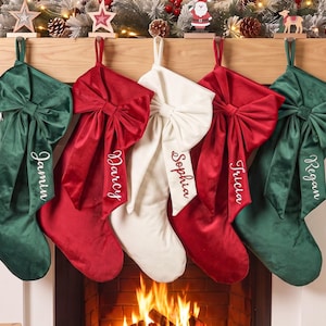 Op de afbeelding: Vijf kerstsokken van fluweel hangen op een rij, bestaande uit twee donkergroene, twee rode en één witte sok. Elke sok is versierd met een grote bijpassende fluwelen strik en gepersonaliseerd met wit cursief borduurwerk met de namen Jamin, Darcy, Sophia, Tricia en Regan.