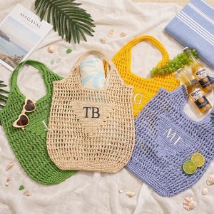 Puede incluir: Cuatro bolsos de mano de ganchillo en verde, beige, amarillo y azul, cada uno con un asa. La bolsa beige tiene las iniciales "TB", la amarilla "G" y la azul "MT". Las bolsas est&aacute;n rodeadas de accesorios de playa.