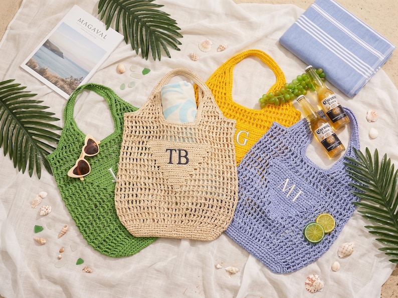 Puede incluir: Cuatro bolsos de mano de ganchillo en verde, beige, amarillo y azul, cada uno con un asa. La bolsa beige tiene las iniciales "TB", la amarilla "G" y la azul "MT". Las bolsas est&aacute;n rodeadas de accesorios de playa.