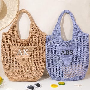 Puede incluir: Dos bolsos tote de tejido abierto, uno beige y otro color lavanda, con iniciales bordadas. El bolso beige tiene bordadas las iniciales "AK", y el bolso lavanda tiene las iniciales "ABS". Un sombrero de paja, gafas de sol y una botella de rosado en el fondo.