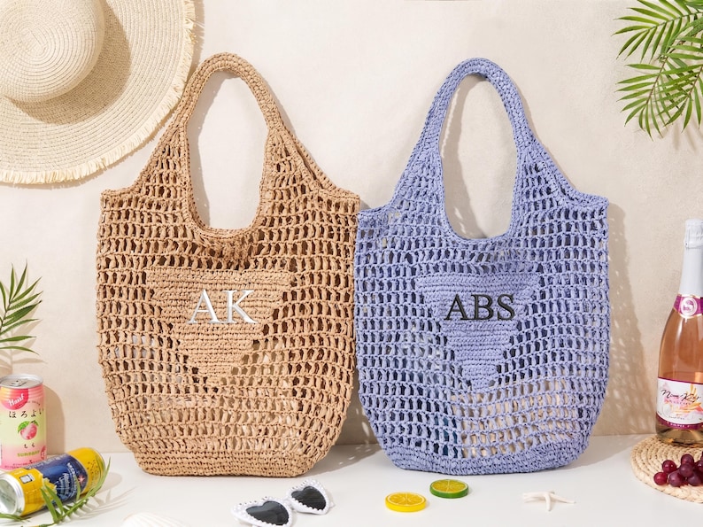 Puede incluir: Dos bolsos tote de tejido abierto, uno beige y otro color lavanda, con iniciales bordadas. El bolso beige tiene bordadas las iniciales "AK", y el bolso lavanda tiene las iniciales "ABS". Un sombrero de paja, gafas de sol y una botella de rosado en el fondo.