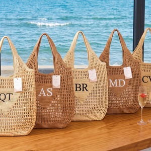 Puede incluir: Cinco bolsos tote tejidos en varios tonos de beige y marrón, cada uno con un diseño de punto abierto y un monograma personalizado de dos letras bordado en negro o blanco. Cada bolso incluye una pequeña etiqueta rectangular colgante.
