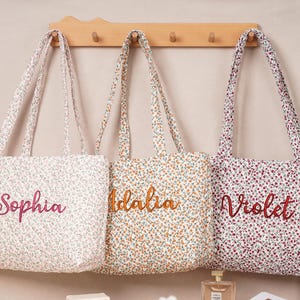 May include: Three floral print tote bags with embroidered names: Sophia, Idalia, and Violet. The bags have matching handles and are displayed against a neutral background. The bags are in shades of white, orange, and purple.