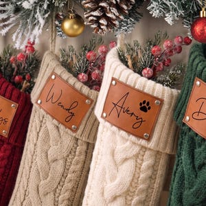 May include: Four cable-knit Christmas stockings in red, beige, cream, and dark green. Each stocking features a rectangular tan leather patch secured with four silver rivets. The patches are personalised with the names Gladys, Wendy, Avery with a paw print, and David in black script.