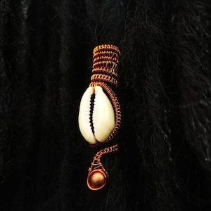 May include: A copper-colored wire hair accessory featuring a white seashell. The wire is intricately wrapped around the shell and forms a spiral design. A small copper bead is at the end of the spiral. The background is black.