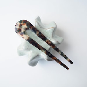 Puede incluir: Una horquilla para el cabello con estampado de carey, con una parte superior en forma de U y dos púas largas y paralelas. El accesorio para el cabello descansa sobre un soporte de cerámica azul claro en forma de estrella sobre un fondo blanco. El patrón incluye marrón, negro y crema.