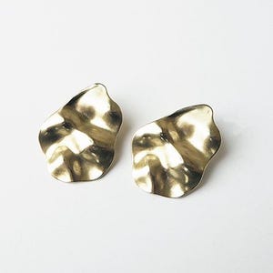 May include: A pair of gold-toned earrings with an irregular, organic, and wavy shape. The surface of each earring features a smooth, reflective metallic finish with gentle indentations that create a rippled, fluid appearance.