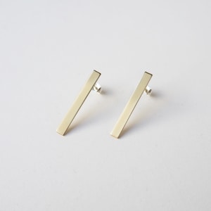 May include: Two gold-coloured bar earrings. Each earring is a straight, rectangular bar with a post backing. The earrings are set against a white background, highlighting their simple, modern design. These earrings are suitable for everyday wear.