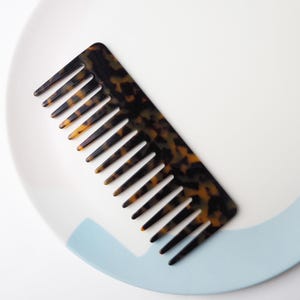 Può includere: Un pettine a denti larghi con un motivo tartarugato, con sfumature di marrone e ambra. Il pettine è posizionato su un piatto bianco con un accento azzurro, offrendo una presentazione pulita ed elegante. Ideale per districare e acconciare i capelli.