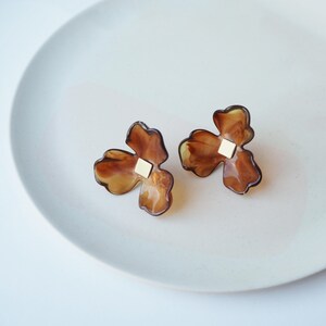 May include: A pair of brown and gold flower-shaped earrings with a square gold centre. The earrings are displayed on a white plate. The earrings have a translucent appearance, with a marbled effect.