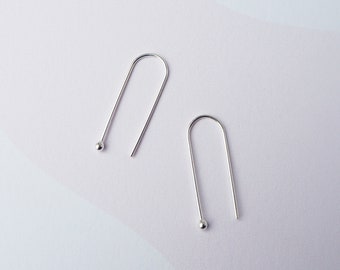 May include: A pair of minimalist silver earrings. Each earring features a curved, elongated design with a small, round ball at the bottom. The earrings are displayed against a white and light purple background.