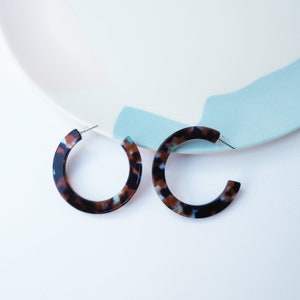 Op de afbeelding: Een paar hoepeloorbellen in schildpadmotief. De oorbellen hebben een gemarmerd patroon van bruin, zwart en lichtblauw. Ze zijn geplaatst tegen een witte achtergrond met een decoratief bord met blauwe en witte accenten.