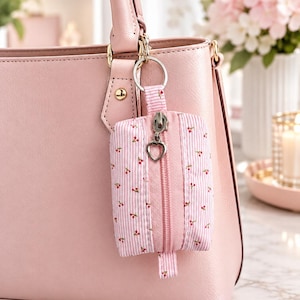 May include: A small rectangular pouch with a pink and white striped pattern featuring tiny red cherry prints. The pouch has a pink zipper closure with a silver heart-shaped pull, attached to a silver keyring hanging from a pink handbag.