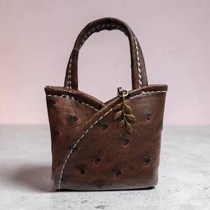 May include: A small brown handbag with a textured, dotted surface pattern, featuring a single arched handle, white contrast stitching along the edges, and a small metallic leaf-shaped charm attached to the front upper rim of the bag.