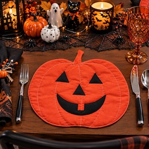 Può includere: Una tovaglietta in tessuto a forma di zucca con un esterno arancione brillante e tratti del viso neri cuciti, inclusi occhi triangolari, un naso triangolare e un'ampia bocca sorridente con un dente, caratterizzata da visibili cuciture bianche.