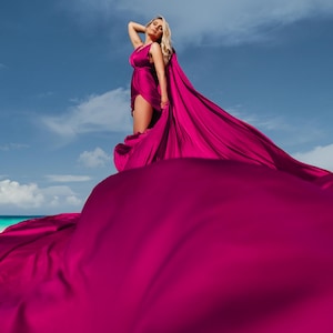 May include: A woman in a magenta dress stands on a beach under a blue sky with clouds. The dress has a flowing train that extends across the sand. The dress is a solid magenta color.