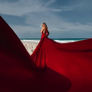 Könnte beinhalten: Eine Frau in einem langen, fließenden roten Kleid steht an einem Strand mit dem Meer im Hintergrund. Das Kleid weht im Wind und erzeugt einen dramatischen Effekt.
