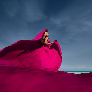 K&ouml;nnte beinhalten: Eine Frau in einem leuchtend pinken Kleid steht an einem Strand, der Wind bl&auml;st den Stoff ihres Kleides hinter ihr her.