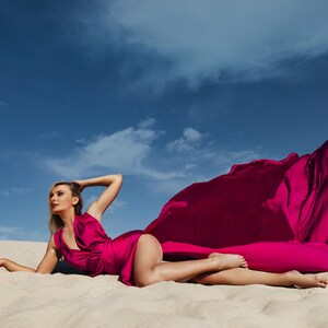 K&ouml;nnte beinhalten: Eine Frau in einem leuchtend pinken Kleid posiert an einem Sandstrand, das Kleid weht im Wind. Der Himmel ist klar blau mit wei&szlig;en Wolken.