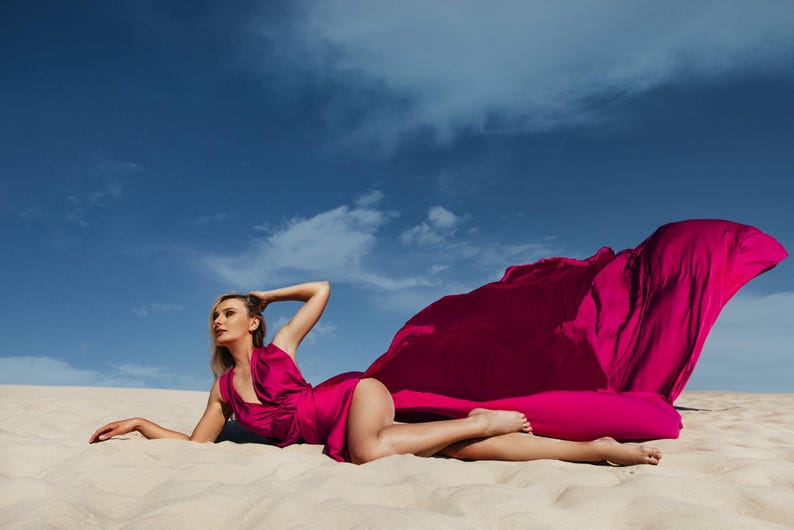 K&ouml;nnte beinhalten: Eine Frau in einem leuchtend pinken Kleid posiert an einem Sandstrand, das Kleid weht im Wind. Der Himmel ist klar blau mit wei&szlig;en Wolken.