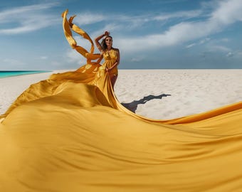 May include: A woman in a yellow dress stands on a white sandy beach with a bright blue ocean in the background. The dress is flowing in the wind, creating a dramatic effect.