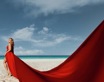 May include: A woman in a flowing red dress stands on a sandy beach with a turquoise ocean and blue sky in the background. The dress has a high slit and is being swept by the wind, creating a dramatic effect.
