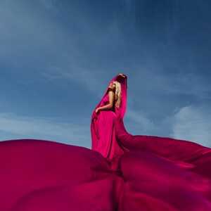 K&ouml;nnte beinhalten: Eine Frau in einem leuchtend pinken Kleid posiert mit dem Stoff, der um sie herum flie&szlig;t und eine dramatische und farbenfrohe Szene vor einem blauen Himmel erzeugt.