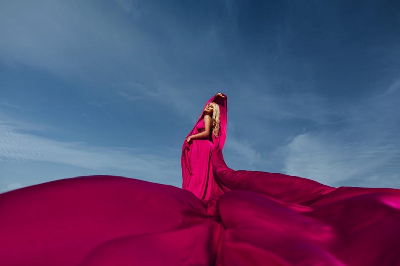 K&ouml;nnte beinhalten: Eine Frau in einem leuchtend pinken Kleid posiert mit dem Stoff, der um sie herum flie&szlig;t und eine dramatische und farbenfrohe Szene vor einem blauen Himmel erzeugt.