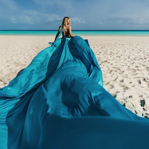 May include: A woman sits on a sandy beach, wearing a flowing teal fabric that trails behind her. The fabric contrasts with the white sand and turquoise ocean under a blue sky. The scene evokes a sense of freedom and elegance.