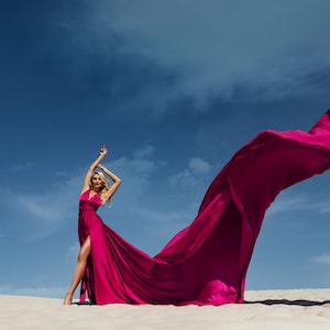 K&ouml;nnte beinhalten: Eine Frau in einem leuchtend pinken Kleid steht an einem Sandstrand, das Kleid weht im Wind. Das Kleid ist lang und flie&szlig;end, mit einem Schlitz an der Seite. Die Frau l&auml;chelt und schaut in die Kamera.