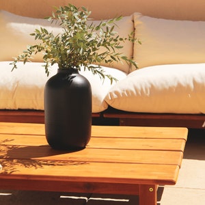 Peut inclure: Un vase en céramique noire avec une plante verte feuillue posé sur une table basse en bois devant un canapé blanc.